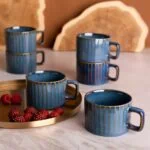 Niebieskie ceramiczne kubki z złotymi paskami na herbatę lub kawę, ustawione na marmurowym blacie z dodatkiem czerwonych malin, idealne do codziennego relaksu i domowego komfortu.