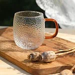 Przezroczysty szklany kubek z pomarańczową rączką na drewnianej desce z makowcami, naturalne światło, rustykalny styl, idealny do napojów, dekoracyjny element domu, wysoka jakość szkła, elegancki i funkcjonalny dodatek kuchenny.