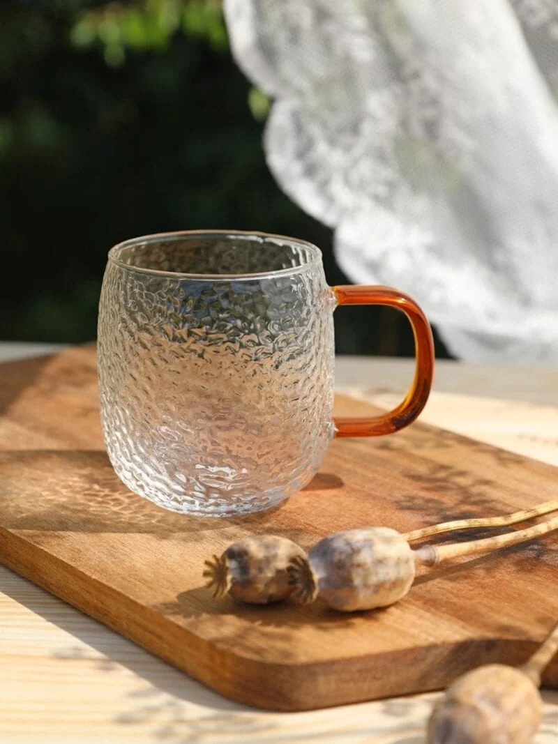 Przezroczysty szklany kubek z pomarańczową rączką na drewnianej desce z makowcami, naturalne światło, rustykalny styl, idealny do napojów, dekoracyjny element domu, wysoka jakość szkła, elegancki i funkcjonalny dodatek kuchenny.