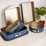 Zestaw tortownic domowych, metalowe foremki w różnych rozmiarach, idealne do pieczenia ciast i serników, nowoczesny design, funkcjonalność, aromatyczne zioła i pomidory na tle białej ściany.