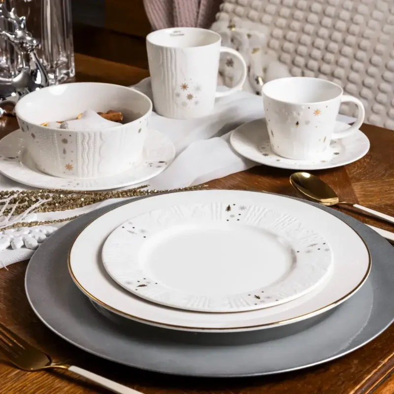 Delikatna, świąteczna zastawa ceramiczna z motywem złotych gwiazdek i śnieżynek, idealna na zimowe i bożonarodzeniowe okazje, elegancko zestawiona na drewnianym stole, w aranżacji Domowego Zakątka.