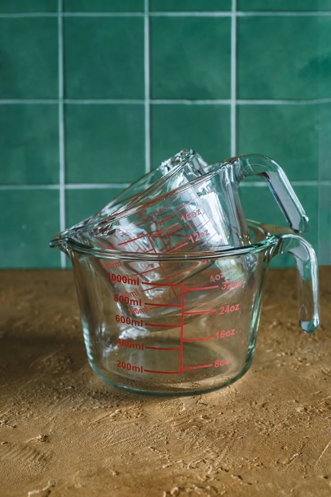 Szklane miarki do pomiaru płynów, zlewki i miarki kuchenne, przechowywanie i odmierzenie składników, nowoczesny design, z zielonym tłem z płytek ceramicznych, praktyczne akcesoria kuchenne, Domowy Zakątek, wysokiej jakości akcesoria kuchenne, wygoda i precyzja w pomiarze, funkcjonalne naczynia do kuchni.
