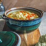 Warzywna zupa w ceramicznym garnku, domowe danie, zdrowa i smaczna, idealna na chłodne dni, w tle kwiaty w wazonie, rustykalny styl kuchni, naturalne składniki, inspiracja do gotowania, kuchnia polska niedroga i pełna smaku, pełnowartościowy posiłek, elegancka zastawa ceramiczna, domowa kuchnia, tradycyjne przepisy, menu na zimę i jesień, odżywcza zupa warzywna, dekoracja stołu, kuchnia w stylu boho, dobrej jakości zastawa, apetyczne danie, zdjęcie ukazujące zdrowe jedzenie, wpisujące się w trendy slow food, domowy sposób na zdrowie, wysokiej jakości ceramika z unikatowym designem, ekologiczne i naturalne składniki, inspiracja do gotowania.