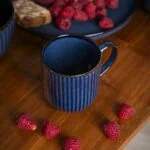 Kubek z ciemnoniebieskiej ceramiki na drewnianym stole, obok świeże maliny i talerz z ciastem, idealny do domowego relaksu i porannej kawy, wpisuje się w tematykę domowego zakątka, smaków i rustykalnego stylu życia.