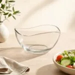 Przezroczysta szklana miska na deser lub sałatkę na jasnym tle, obok talerz z warzywami, minimalistyczny wystrój kuchni, naturalne światło, produkt z oferty sklepu domowyzakatek.pl.