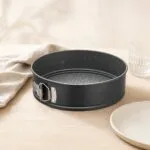 Okrągła forma do pieczenia z nieprzywierającą powłoką, idealna do ciast i tart, czarna, na drewnianym blacie z szklanką i talerzem w tle.