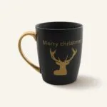 Miedziany uchwyt i złoty napis "Merry Christmas" na czarnym kubku ceramicznym z rysunkiem renifera, idealny na świąteczne okazje, element dekoracji kuchni i prezent świąteczny.