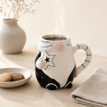 Przedstawienie kubka z ceramiki w kształcie postaci z uśmiechniętą twarzą, z czarnymi i różowymi detalami, na jasnym stole z talerzem ciastek i kwiatami w tle, idealne do kawy lub herbaty.