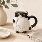 Szczotkowana ceramiczna filiżanka w kształcie psa z czarnym uchem i ogonem, idealna na kawę lub herbatę, na tle jasnego stołu i dekoracyjnej doniczki z zielonymi liśćmi.