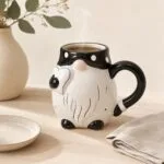 Szczotkowana ceramiczna filiżanka w kształcie psa z czarnym uchem i ogonem, idealna na kawę lub herbatę, na tle jasnego stołu i dekoracyjnej doniczki z zielonymi liśćmi.