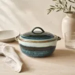 Talerz ceramiczny z pokrywką w odcieniach niebieskiego i białego, na jasnym tle, obok szklanka i wazon z gałązkami, idealny do kuchni i serwowania potraw.