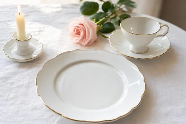 Klasyczny prezent na Komunie - bialy porcelanowy talerz ze zlotym rantem, filizanka, swiecznik z swieca i rozowa roza na bialym lnianym obrusie